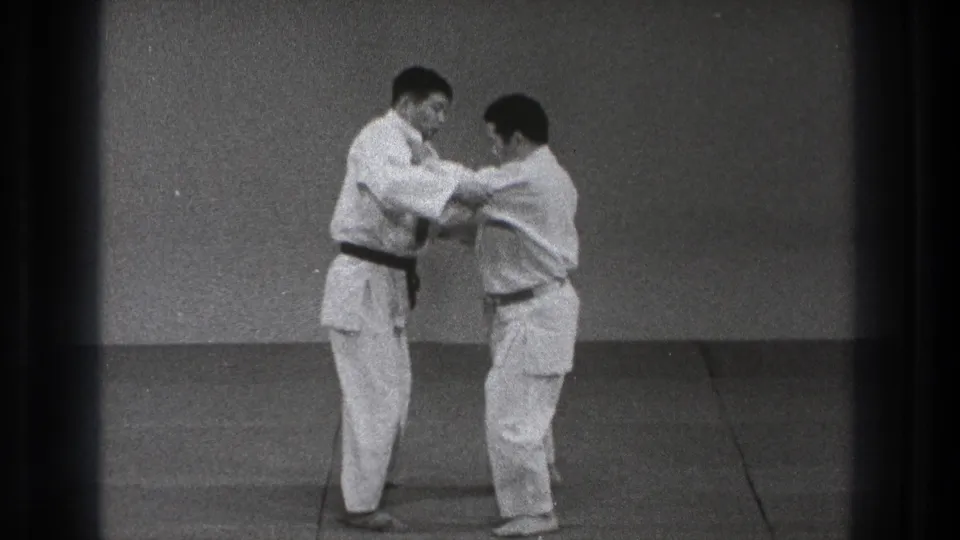 Two adult judo practitioners continue a close grappling technique on the mat, with one man wearing a black belt.