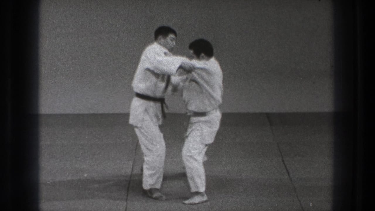 In Tokyo 1971 footage, two adult men in white judo uniforms begin a paired technique on a plain indoor mat.