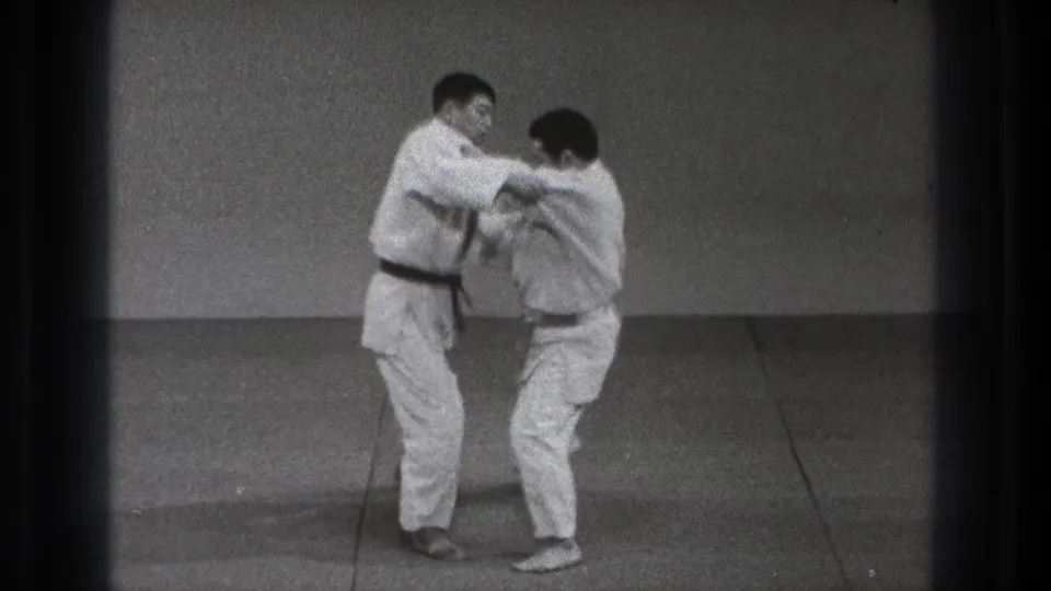 In Tokyo 1971 footage, two adult men in white judo uniforms begin a paired technique on a plain indoor mat.