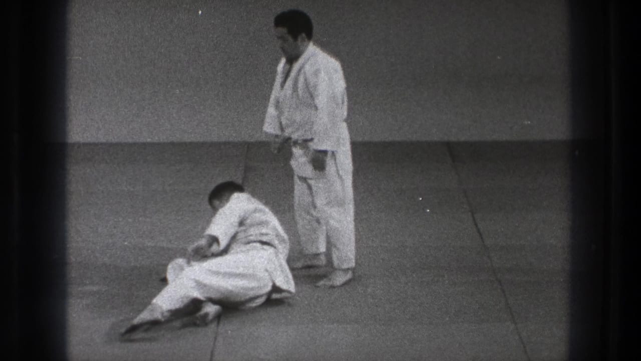 A 1971 Tokyo judo practice moment shows one adult man seated after the fall while his partner stands beside him.