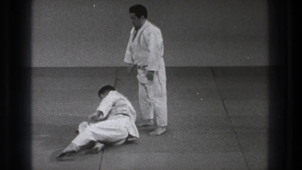 A 1971 Tokyo judo practice moment shows one adult man seated after the fall while his partner stands beside him.