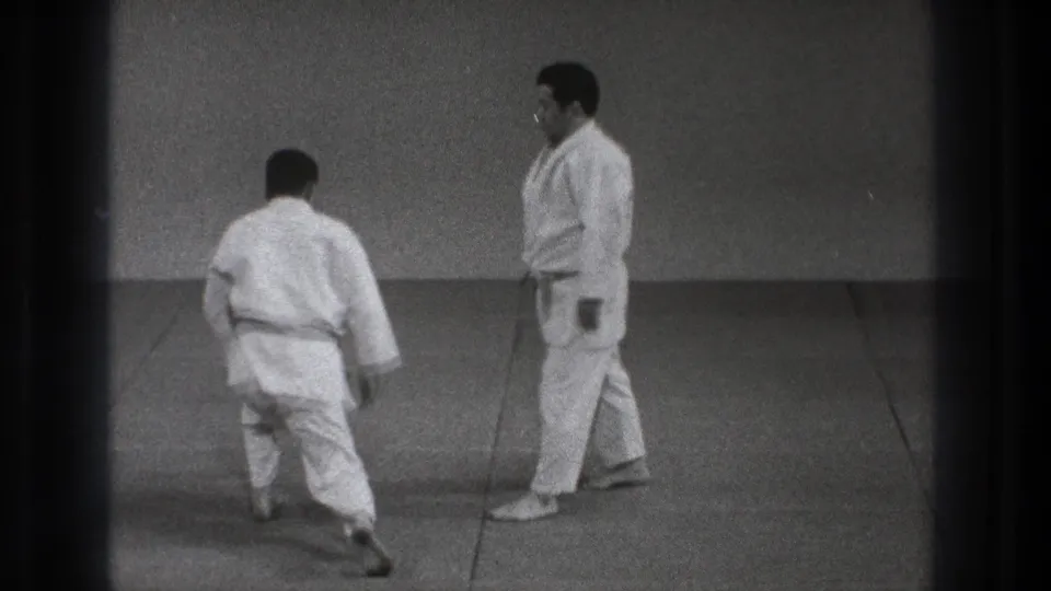 Two men in white judogi appear on a Tokyo dojo mat in 1971, one walking away as the other stands nearby.