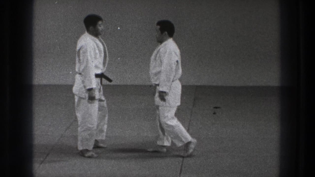 Two judo practitioners in white uniforms face one another on a mat, with a black belt visible on the left figure.