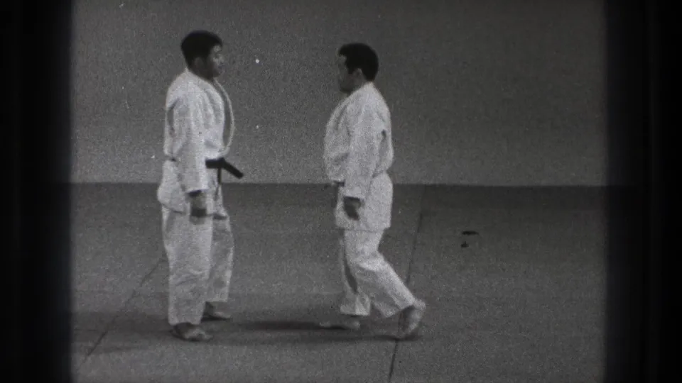 Two judo practitioners in white uniforms face one another on a mat, with a black belt visible on the left figure.
