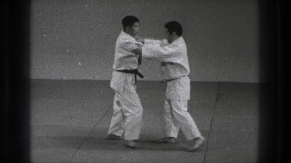Two judo practitioners in white uniforms begin a standing grip on the mat, with rank belts visible.