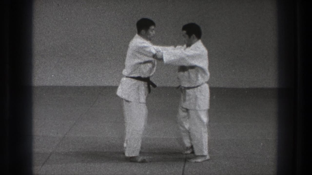 Two men in white gi grip each other on a plain indoor mat.