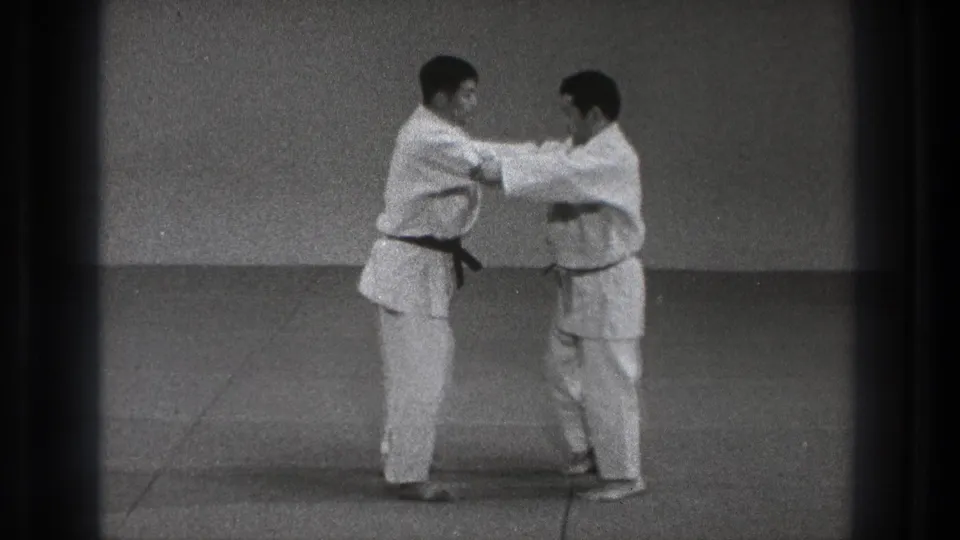In 1971 Tokyo footage, two adult men in white martial arts uniforms begin a controlled grip exchange on an indoor mat.