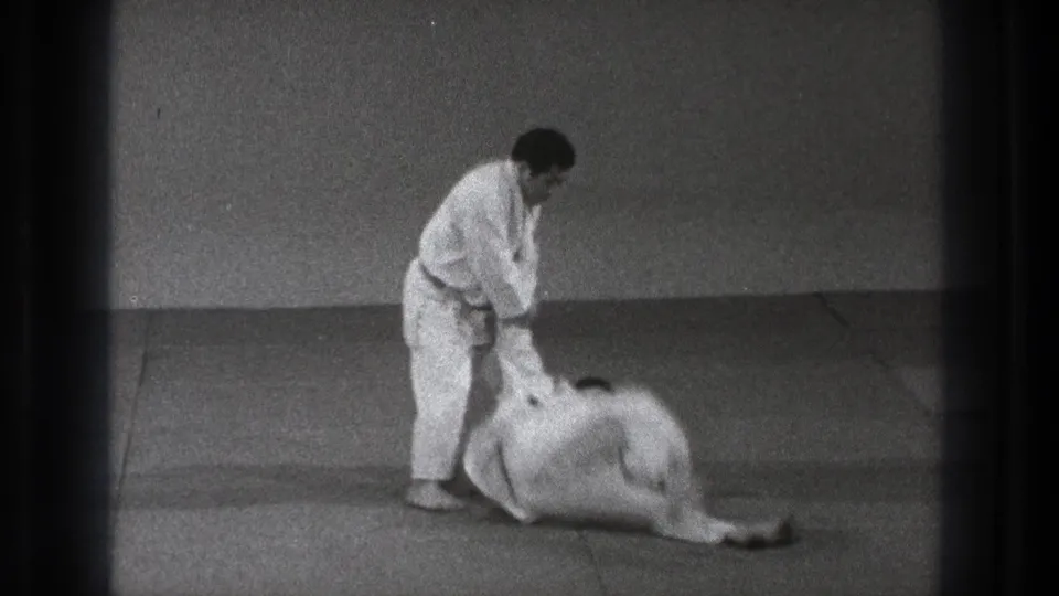 During the 1971 demonstration, one practitioner goes down toward the mat as the standing man maintains arm control.