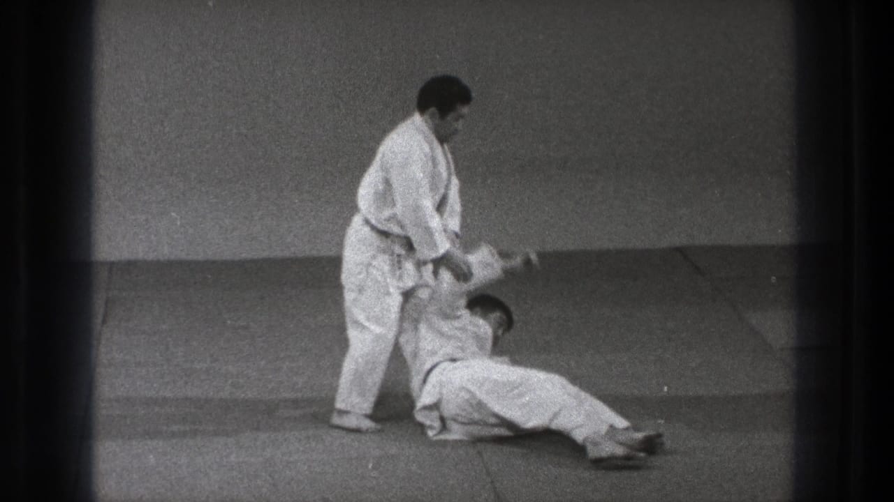 The technique ends with one practitioner seated on the mat and the standing black-belt practitioner holding his arm.