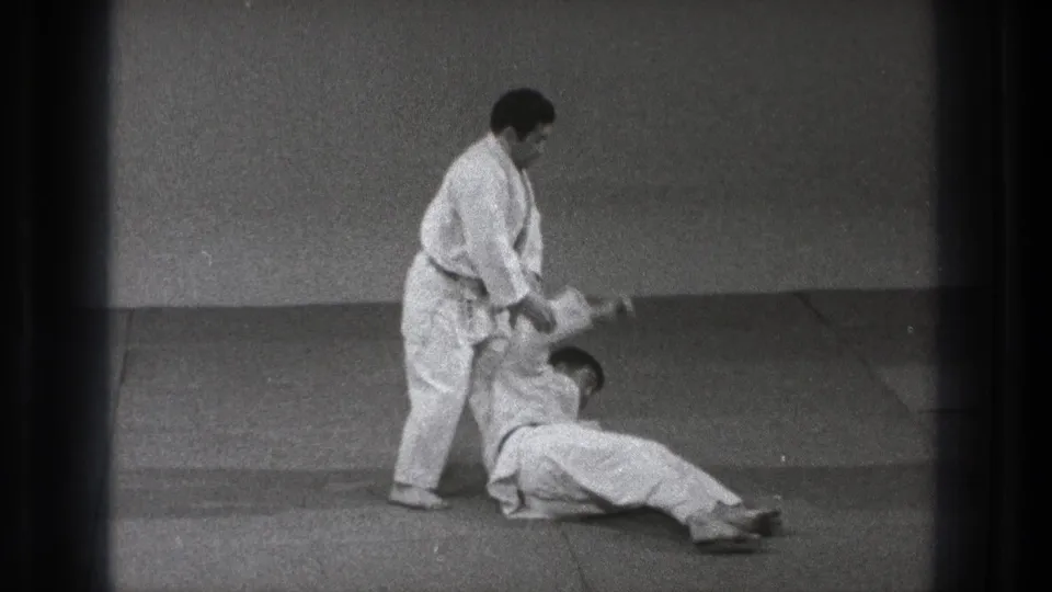 The technique ends with one practitioner seated on the mat and the standing black-belt practitioner holding his arm.