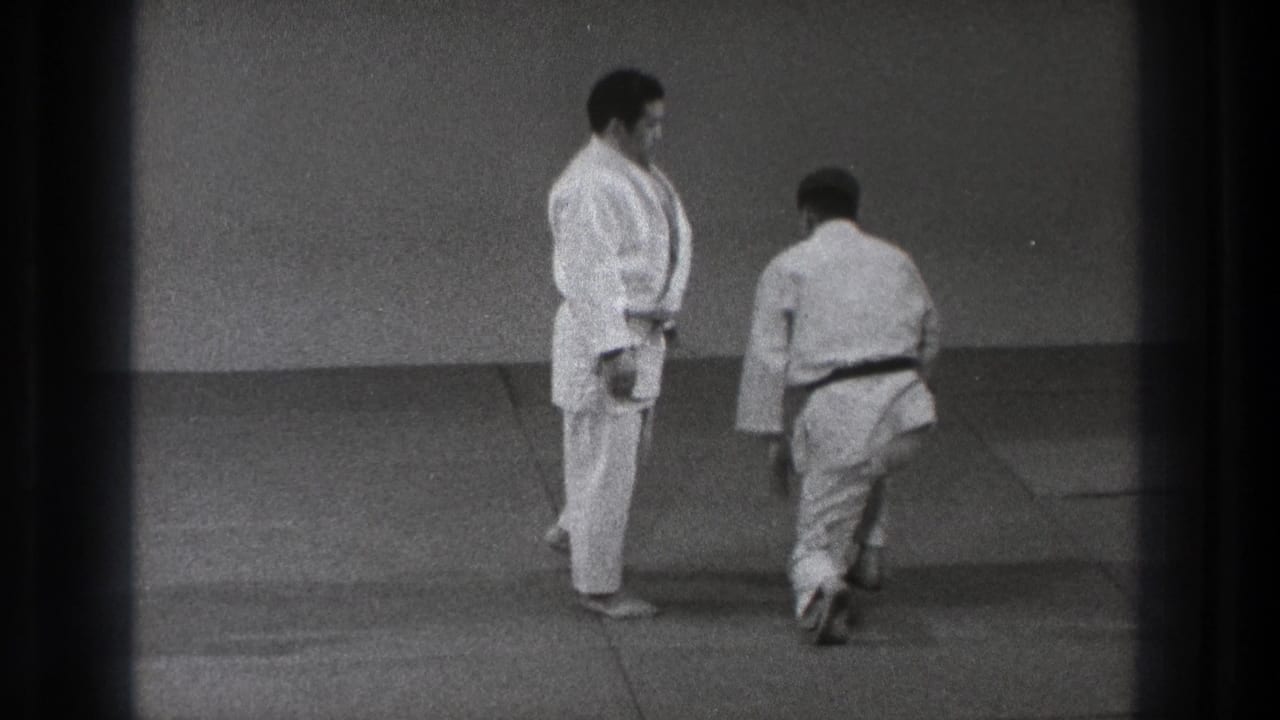 In the Tokyo training room, one gi-clad practitioner steps away while a dark-belted partner watches on the mats.