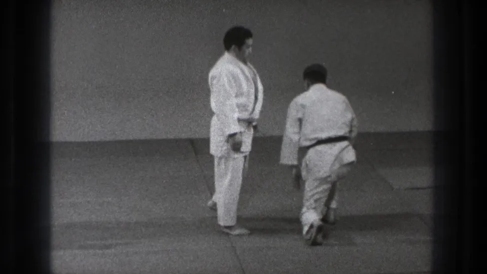 In the Tokyo training room, one gi-clad practitioner steps away while a dark-belted partner watches on the mats.