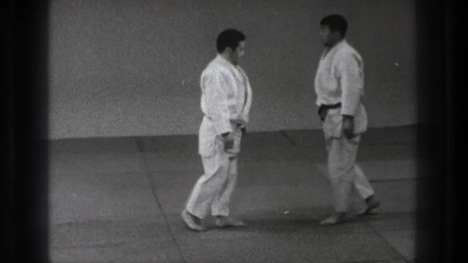 Two adult martial arts practitioners in white gi face each other on mats in the supplied 1971 Tokyo footage.