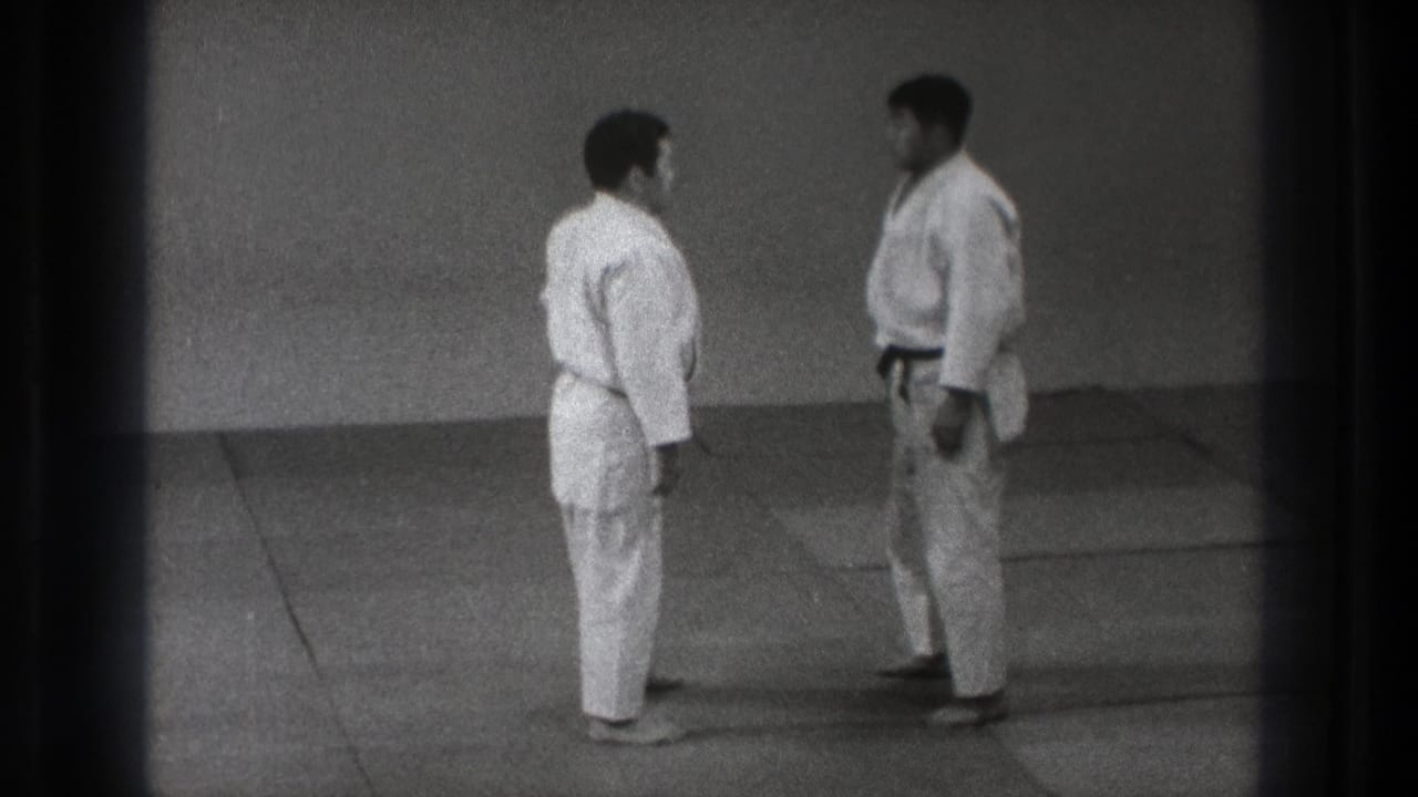 The martial arts pair stands face to face on the mat, framed in grainy black-and-white 8mm footage from Tokyo.