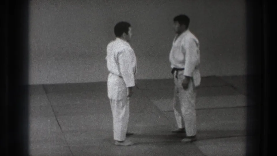 The martial arts pair stands face to face on the mat, framed in grainy black-and-white 8mm footage from Tokyo.