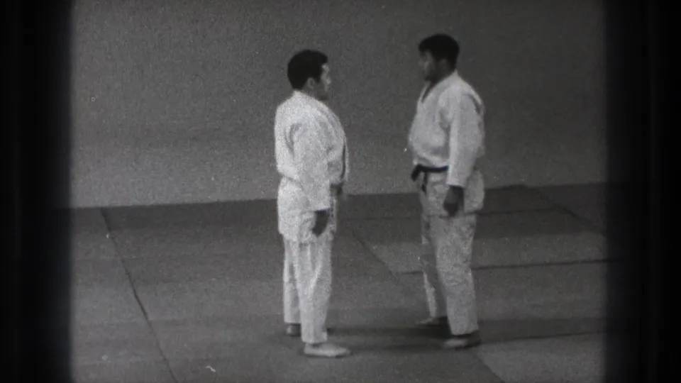 In Tokyo, Japan in 1971, two adult men in white judo gis and dark belts stand facing each other on tatami mats.