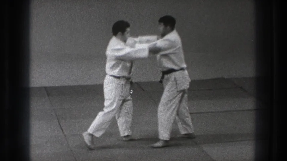 Two adult men in judo gis make sleeve and lapel contact on tatami mats during a 1971 Tokyo practice exchange.