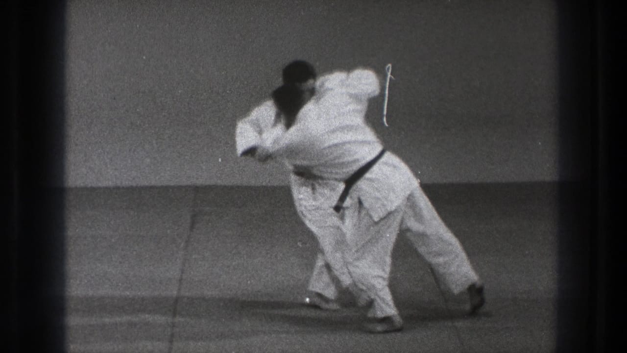 The judo exchange reaches the leg take-down moment, with one man turning and pulling the other off balance on the tatami.