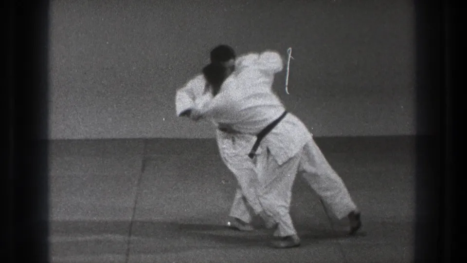 The judo exchange reaches the leg take-down moment, with one man turning and pulling the other off balance on the tatami.