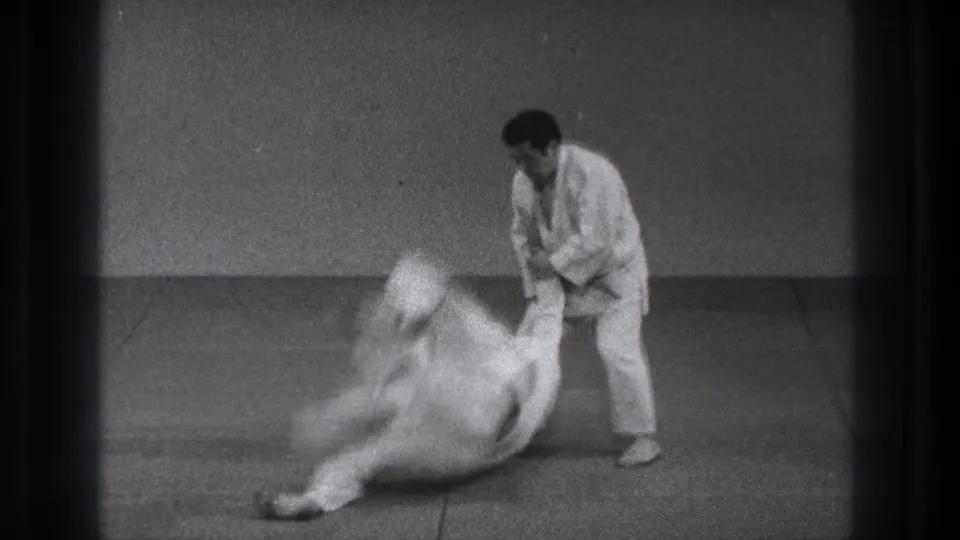 1971 Tokyo judo practice frame showing two adult men in white judogi as a black-belt practitioner initiates a throw on tatami.