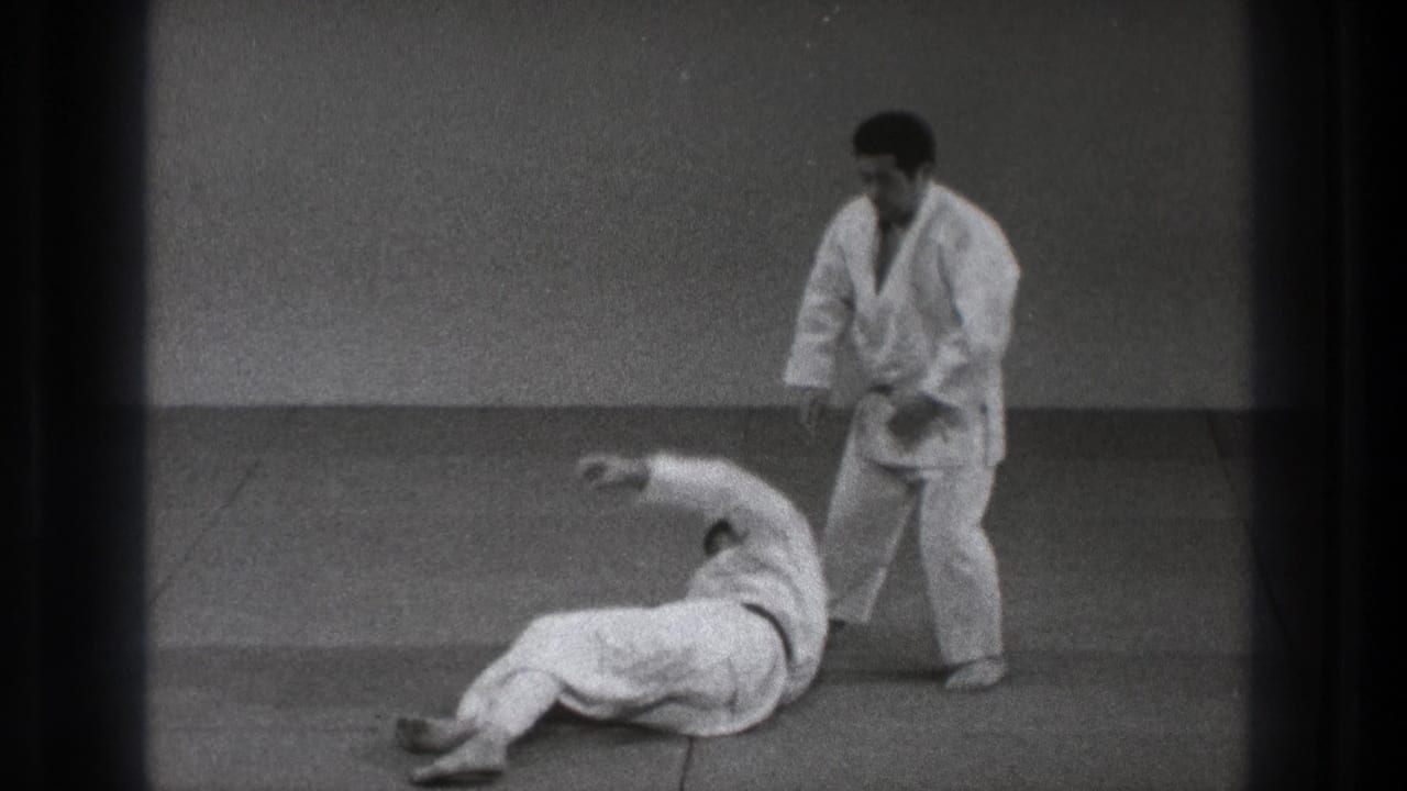 1971 Tokyo judo practice frame with one adult man on the tatami after a throw and the black-belt practitioner standing beside him.