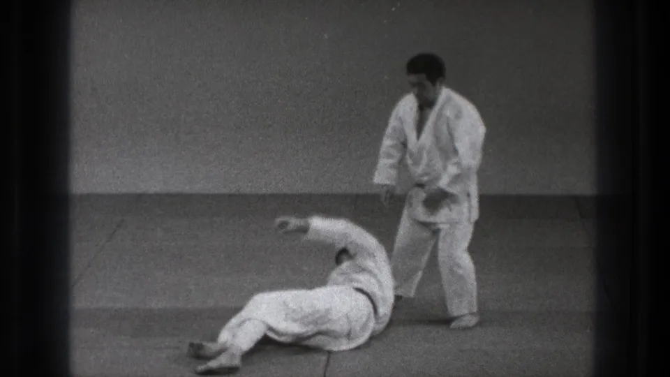 1971 Tokyo judo practice frame with one adult man on the tatami after a throw and the black-belt practitioner standing beside him.