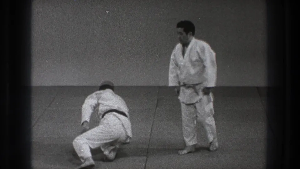 1971 Tokyo judo practice frame showing one adult practitioner kneeling after the throw while the black-belt partner stands on the tatami.