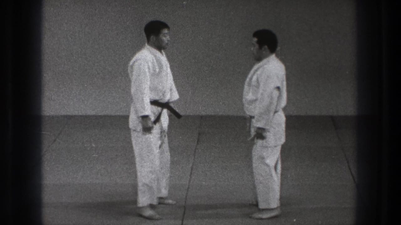 Two adult karate practitioners in white gi remain squared off on the dojo mat, with the black belt visible on the taller man.