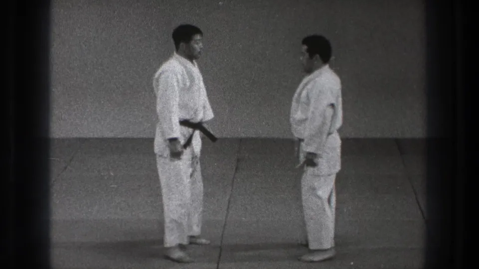 Two adult karate practitioners in white gi remain squared off on the dojo mat, with the black belt visible on the taller man.