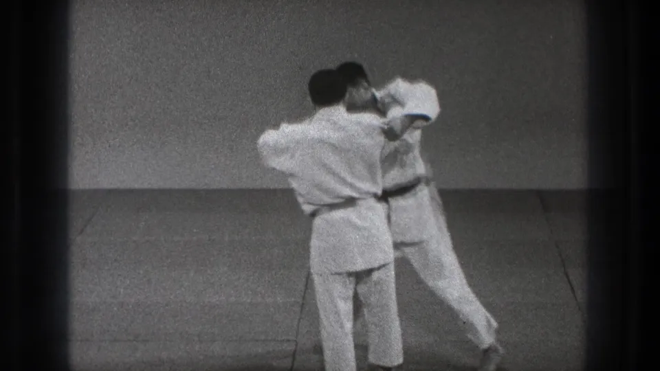 In 1971 Tokyo footage, two adult judo practitioners in white gi begin a standing grip exchange on a practice mat.