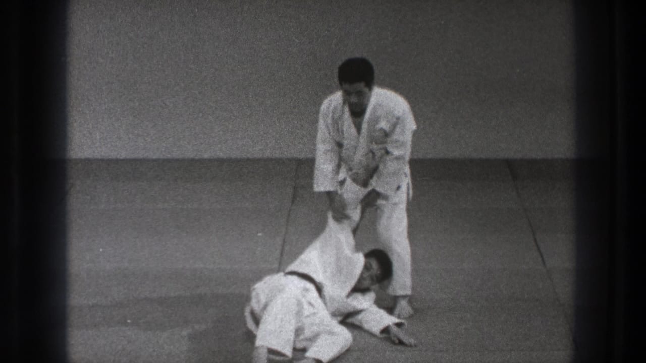 Two men in white martial arts uniforms practice a throw on a mat.