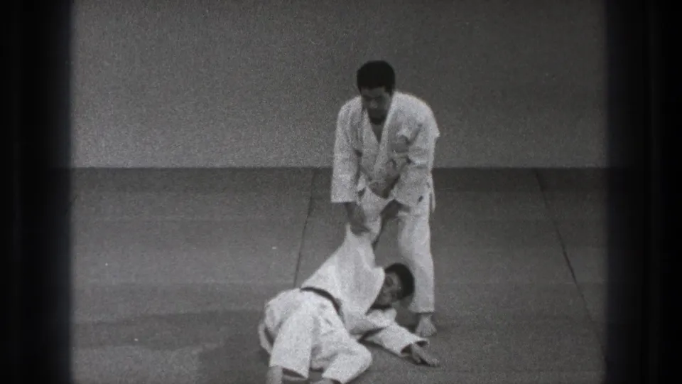 In 1971 Tokyo footage, one man in a white gi stands over another man on the mat during a martial arts technique.