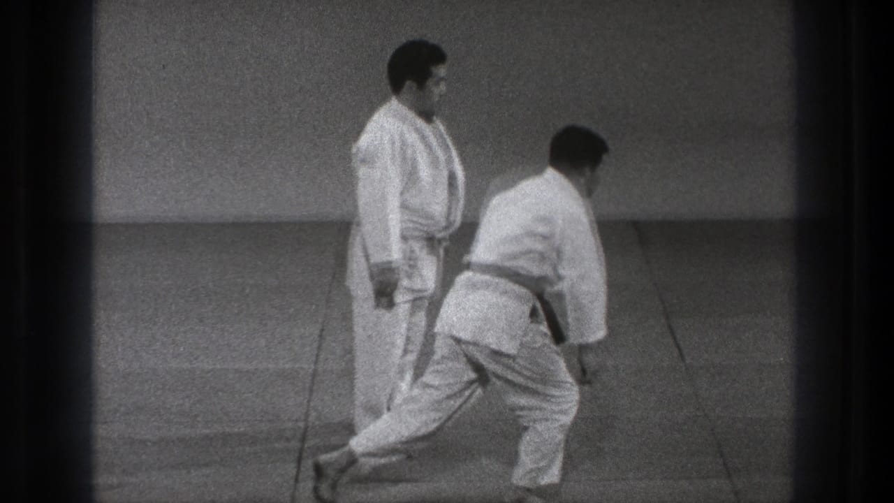 The two adult practitioners reset in the sparse Tokyo training space, with one stepping forward and the other watching.