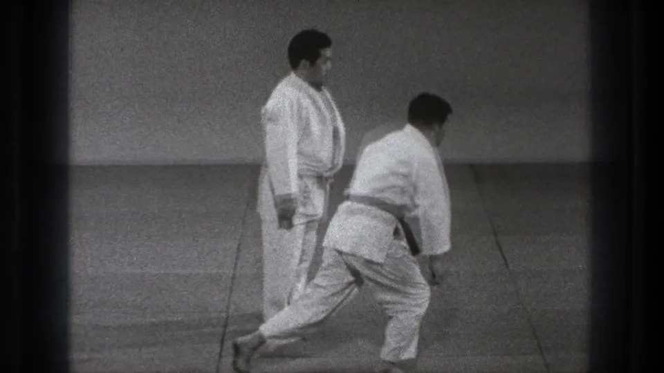 The two adult practitioners reset in the sparse Tokyo training space, with one stepping forward and the other watching.