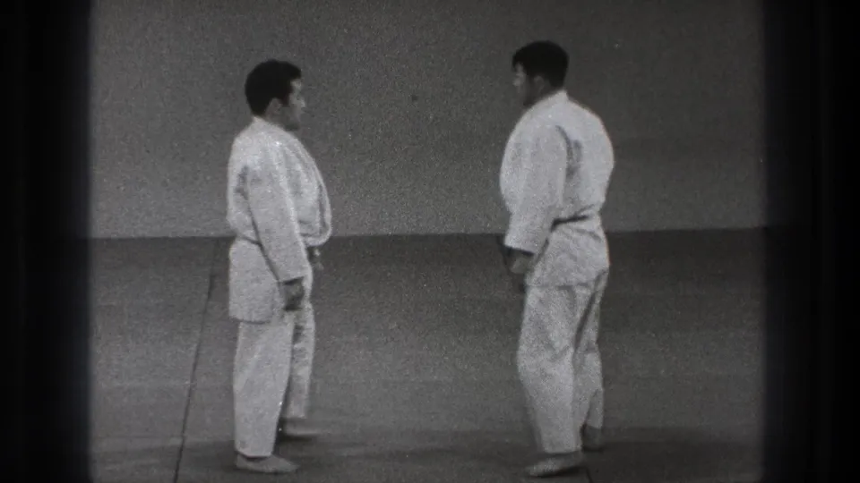 In 1971 Tokyo footage, two adult men in white gi face each other on a training mat before beginning a judo exchange.