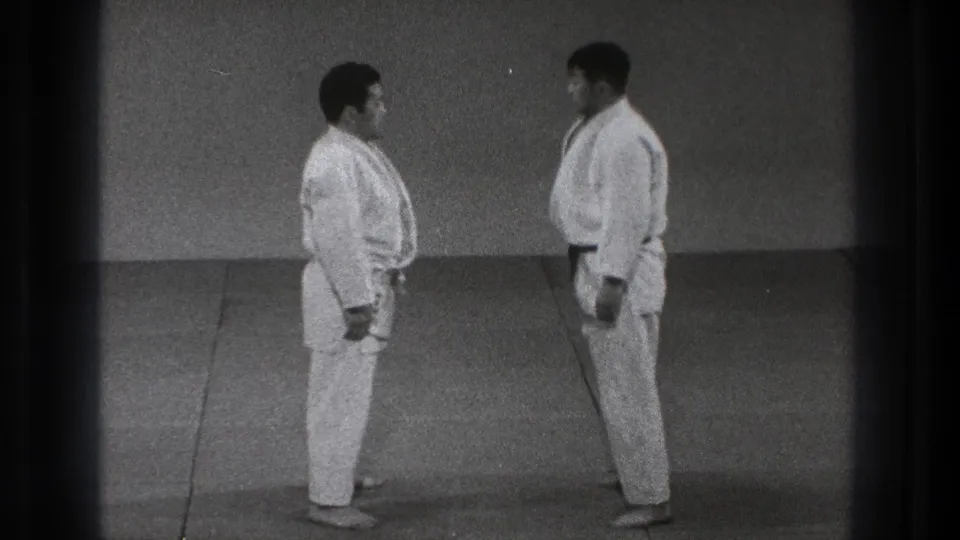 Two men in white gi stand in profile on a matted floor, part of a 1971 Tokyo home-movie judo practice scene.