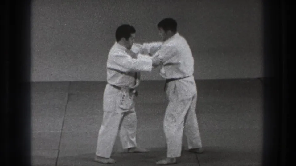 In the 1971 Tokyo judo practice clip, two men grip each other's gi while preparing a controlled technique on the mat.