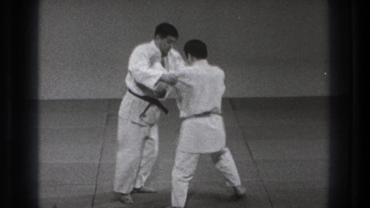 Two men in white gi begin a karate defense drill on a mat.