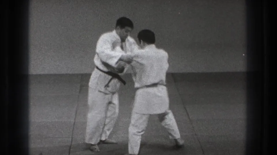 In 1971 Tokyo footage, two adult men in white gi face each other on a mat as a black-belt instructor controls his partner's arm.