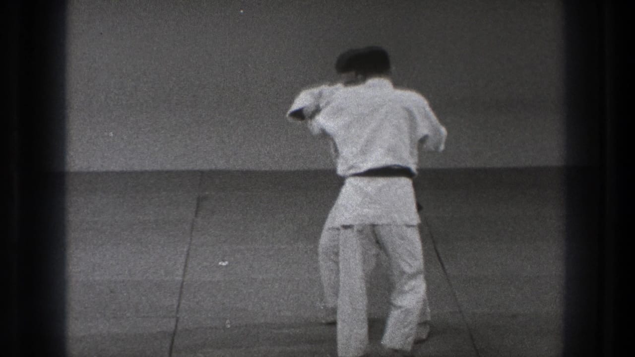The karate pair remains standing as the black-belt practitioner turns into a close grip during the defensive sequence.