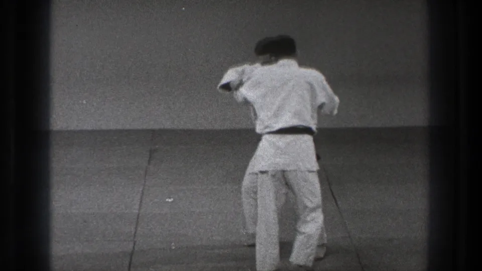 The karate pair remains standing as the black-belt practitioner turns into a close grip during the defensive sequence.