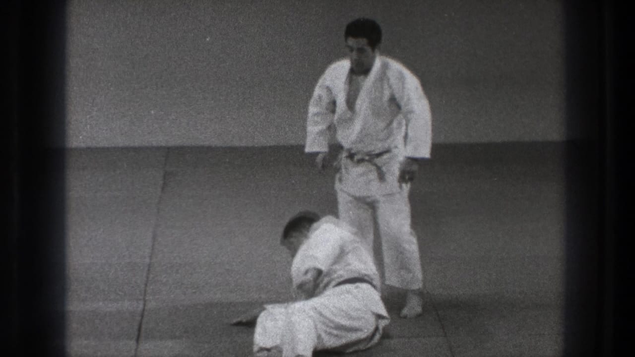 1971 Tokyo 8mm frame showing two adult men in judo uniforms on a padded mat, with one seated low and the other standing in observation.