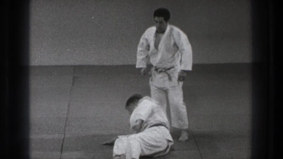 1971 Tokyo 8mm frame showing two adult men in judo uniforms on a padded mat, with one seated low and the other standing in observation.