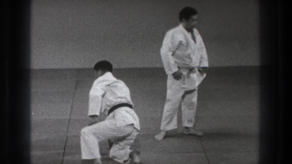 1971 Tokyo practice frame with one man getting up from the mat and another adult judo practitioner standing nearby in a plain indoor space.