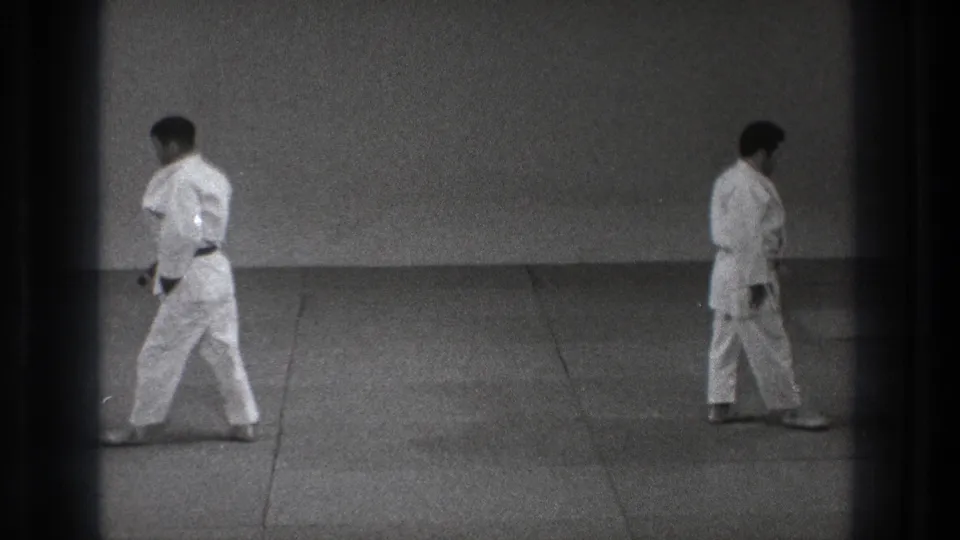 1971 Tokyo 8mm frame with two adult men in white judo uniforms walking apart across a padded mat after the practice sequence.