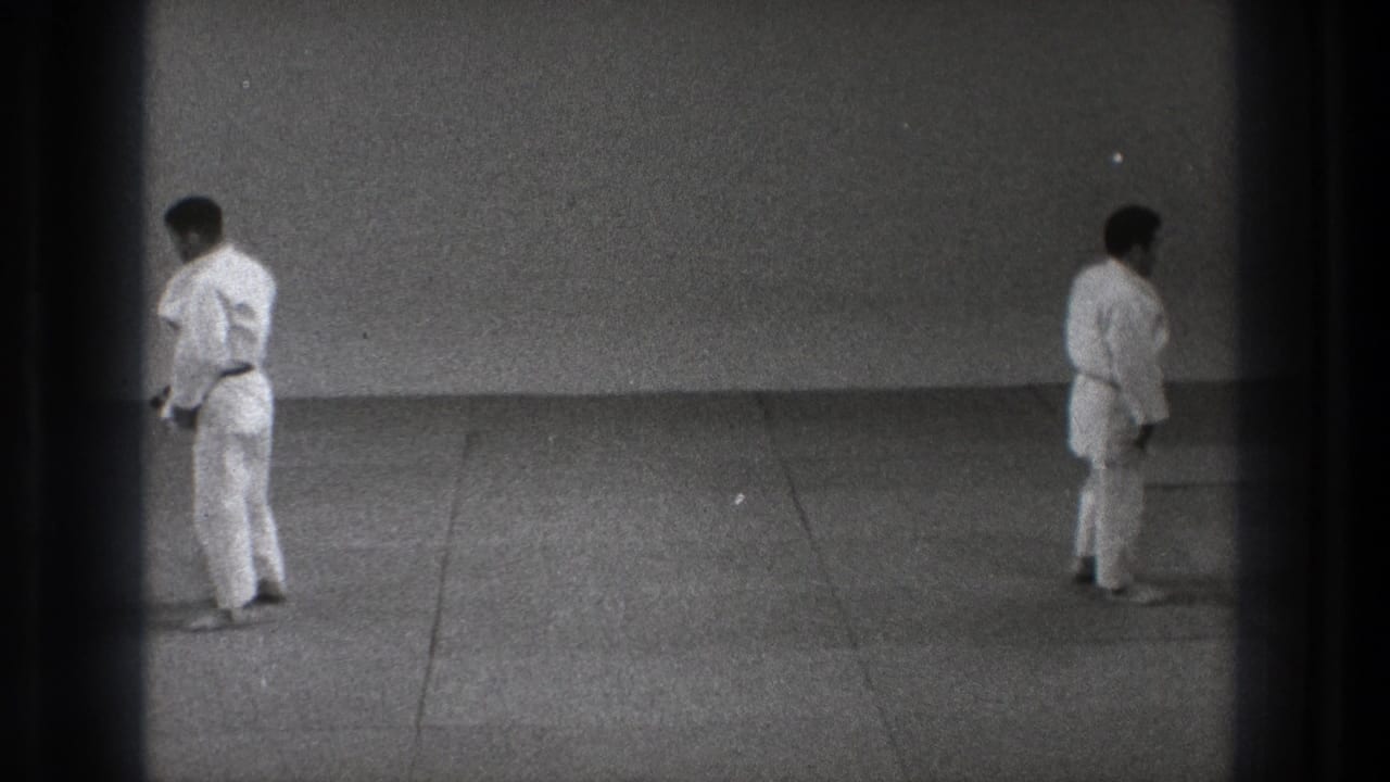 In supported 1971 Tokyo footage, two adult men in karate gi face each other across a tatami mat in a plain practice room.