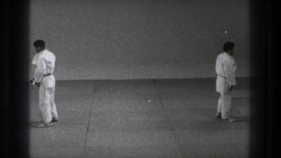 In supported 1971 Tokyo footage, two adult men in karate gi face each other across a tatami mat in a plain practice room.