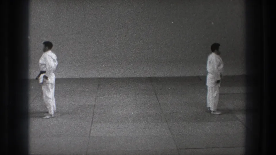 Two adult karate practitioners in white gi stand in profile on a tatami mat, with one dark belt visible against the plain dojo wall.