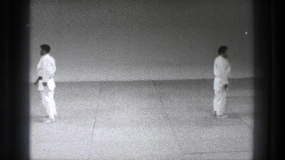 The 8mm frame shows two adult men in karate gi spaced across a tatami mat, viewed from a fixed home-movie perspective.
