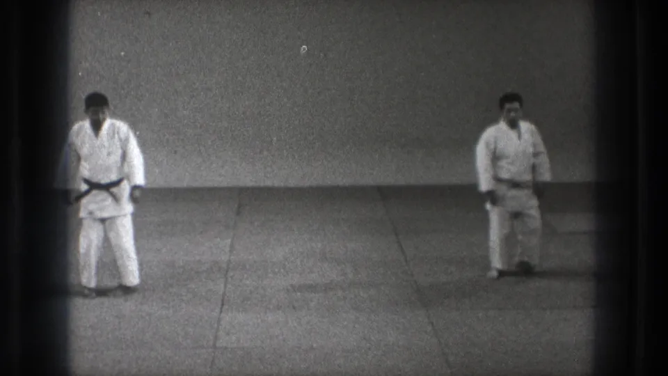 Black-and-white 8mm frame of two adult judoka in white uniforms spaced across a tatami mat during a demonstration setup.